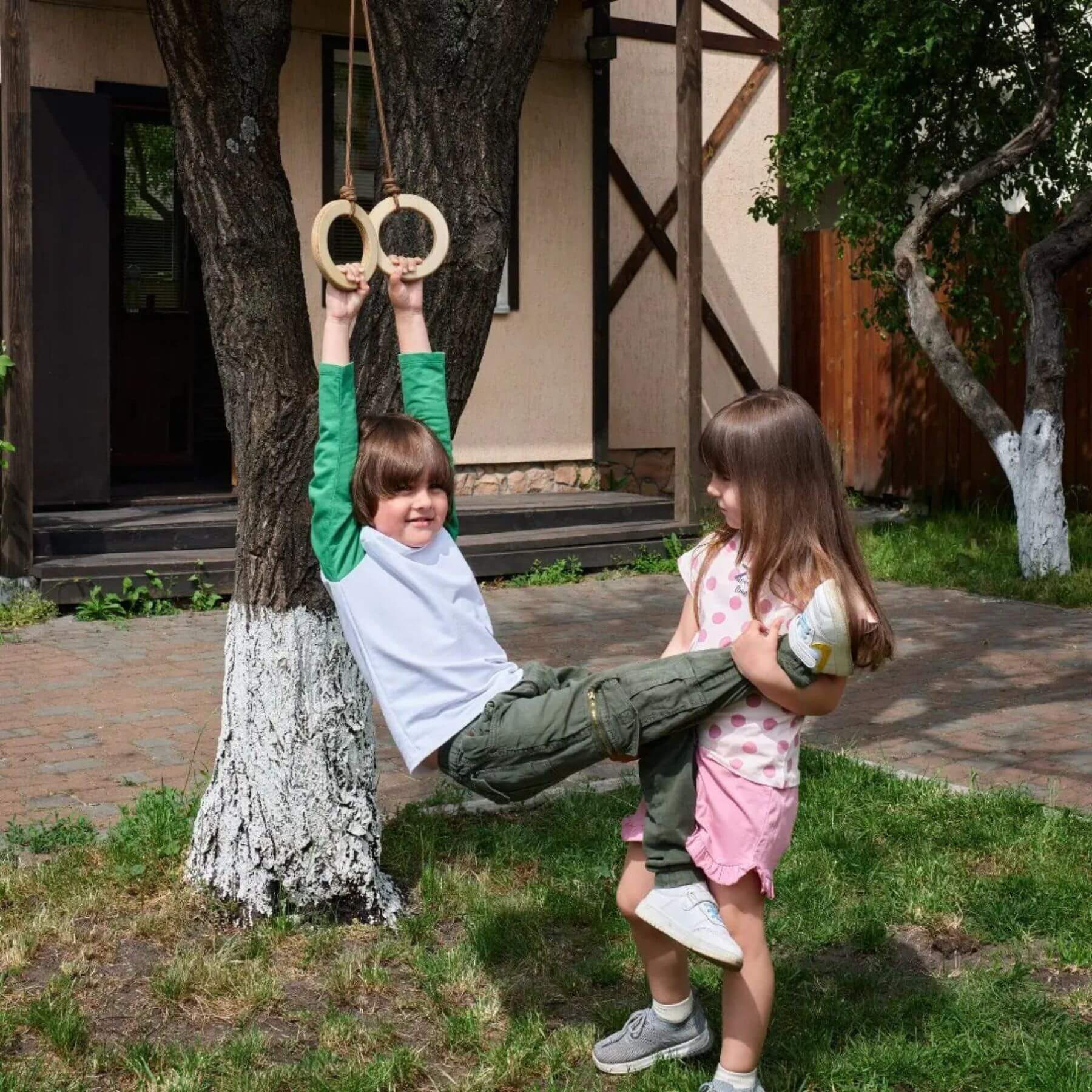Two children playing with rings on a tree outside a house.