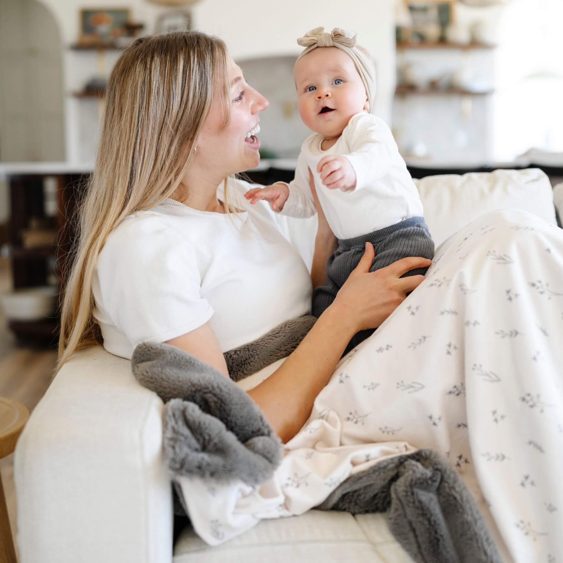 Woman holding a baby on a couch covered by Saranoni Minky/Lush Throw Blankets - Jasmine in a cozy living room.