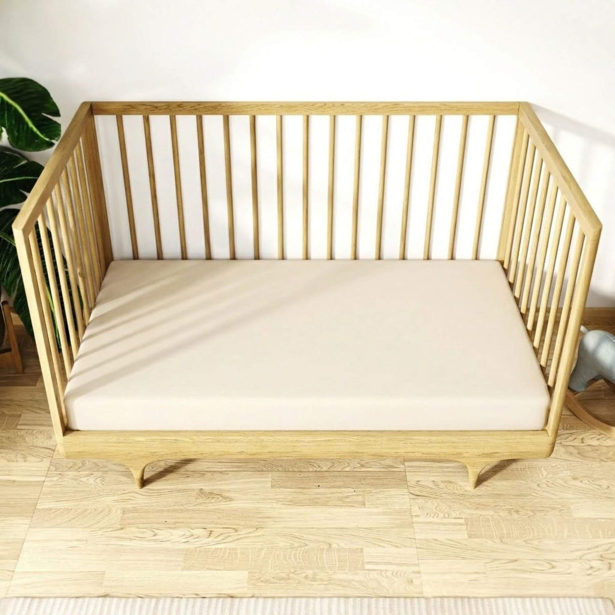 A light natural wood crib or daybed with vertical slat sides and a cream-colored mattress, shown in a bright room with light wood flooring and a white wall.