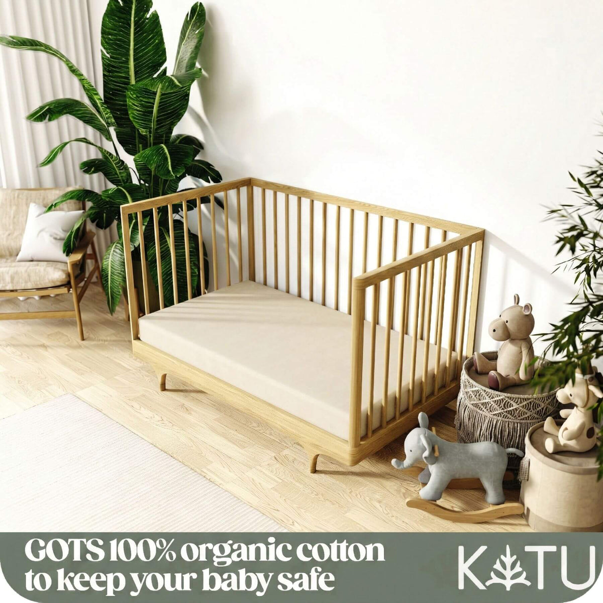 A light natural wood baby crib with a cream mattress, styled in a bright nursery with a large green plant, a rocking horse, and toys. A dark green banner at the bottom features the text 'GOTS 100% organic cotton to keep your baby safe' and the brand logo 'KATU'.