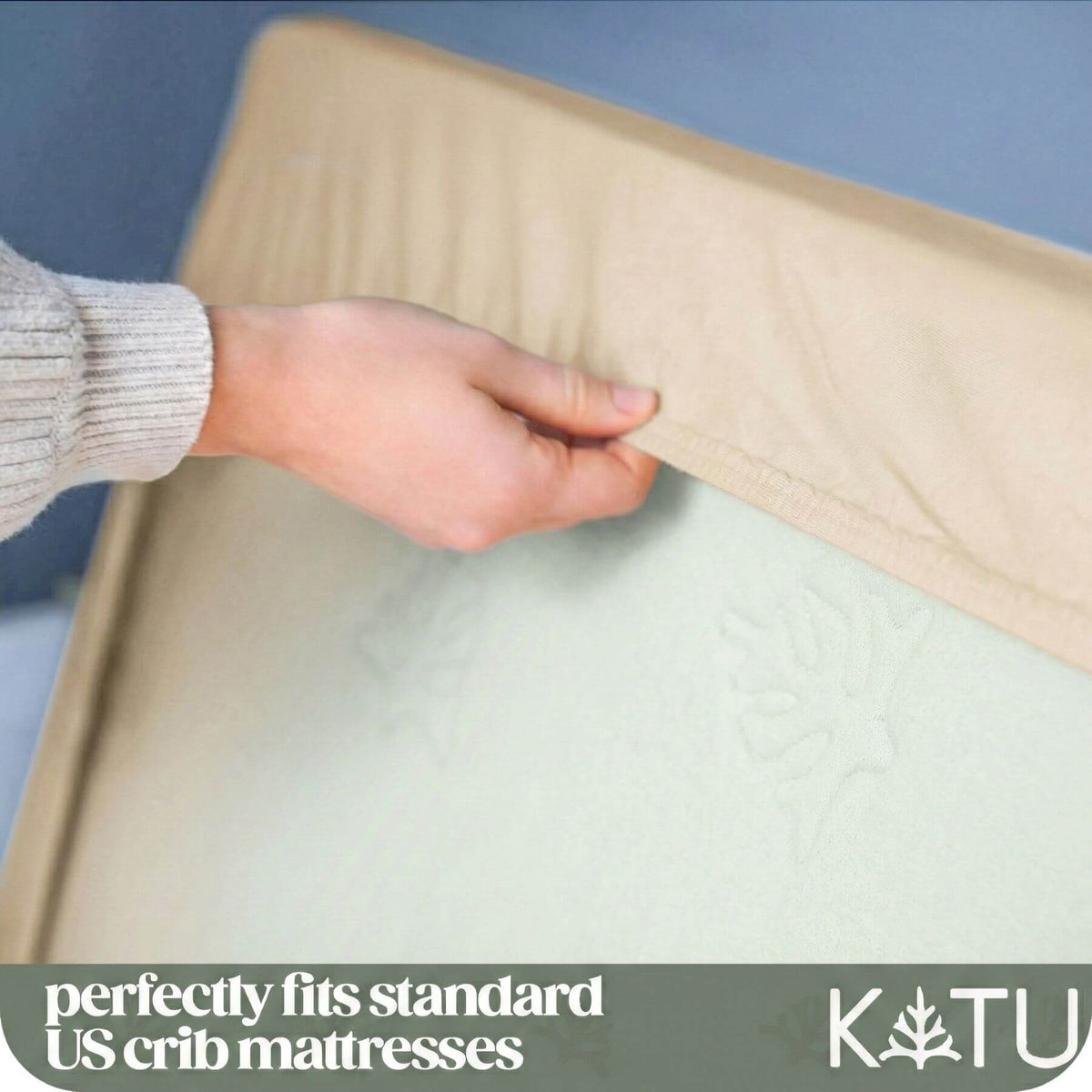 A close-up of a hand lifting the fitted sheet off a mattress to show the mattress fit, with text at the bottom reading 'perfectly fits standard US crib mattresses' and a K&U brand mark.