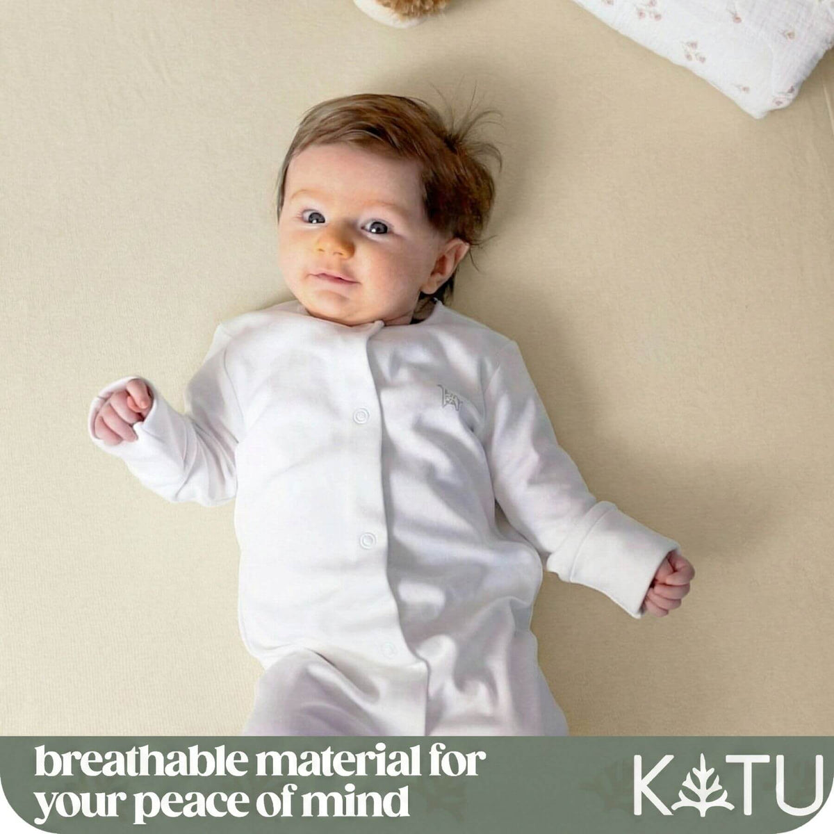 A baby lying on a beige blanket wearing a white long-sleeve sleep sack. A text banner at the bottom reads 'breathable material for your peace of mind' with the brand name 'KATU' on the right.