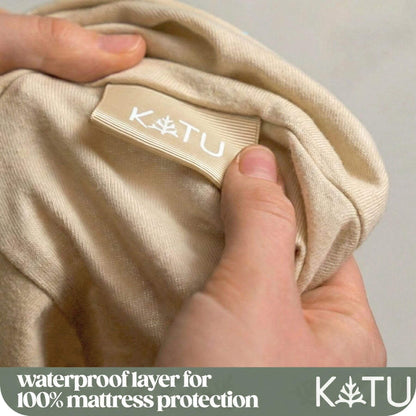 Close-up of a person's hands holding a beige fabric item to show a sewn-on KATU label, with a text banner at the bottom reading 'waterproof layer for 100% mattress protection' and the KATU logo.