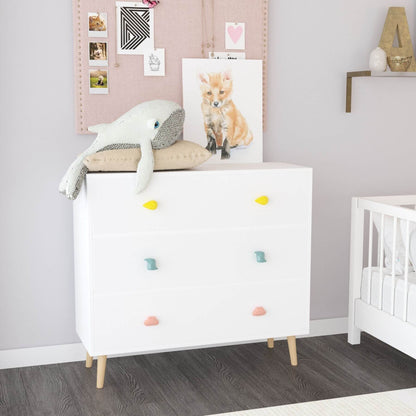 White dresser with colorful knobs in a nursery setting with a crib and wall decorations.