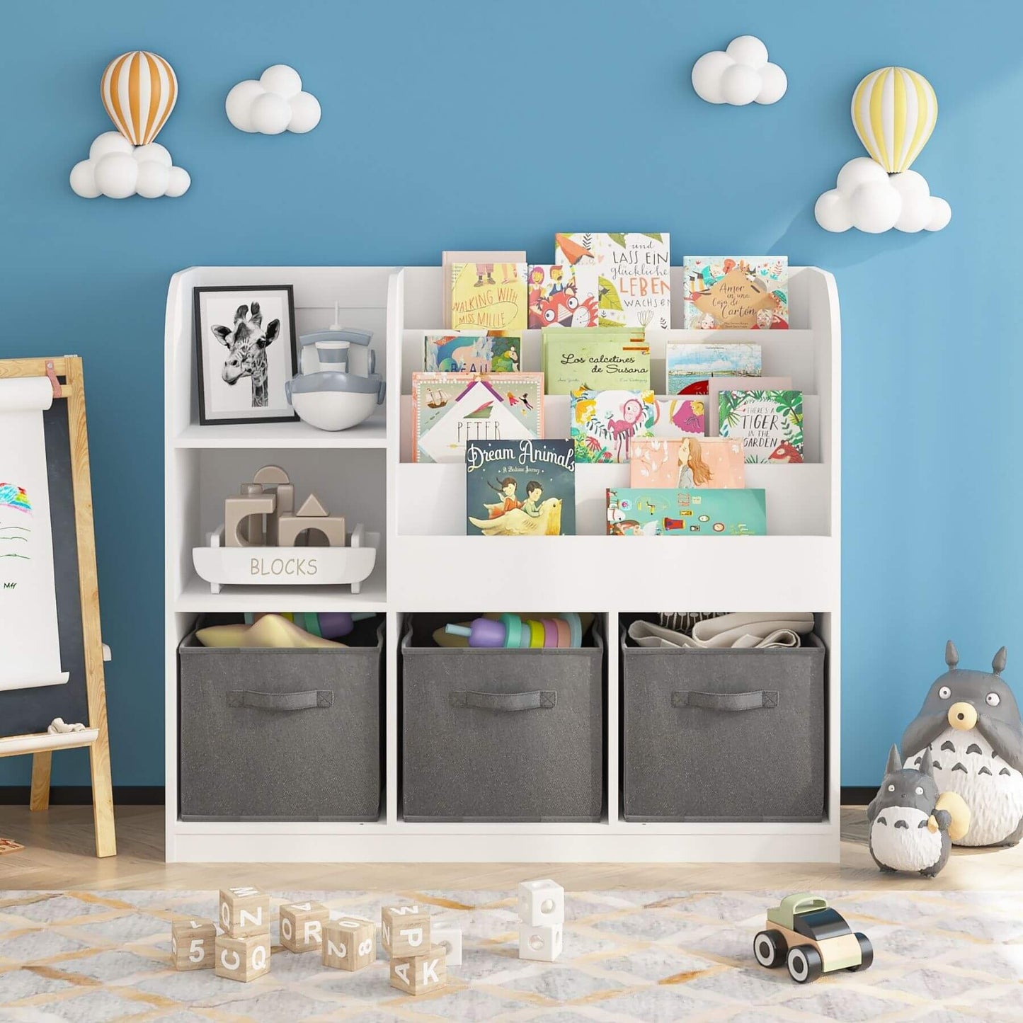 Children's room with MOONRIVER Kids Bookcase with 3 Collapsible Fabric Drawers - White/Gray filled with books and toys against a blue wall with cloud and balloon decorations.