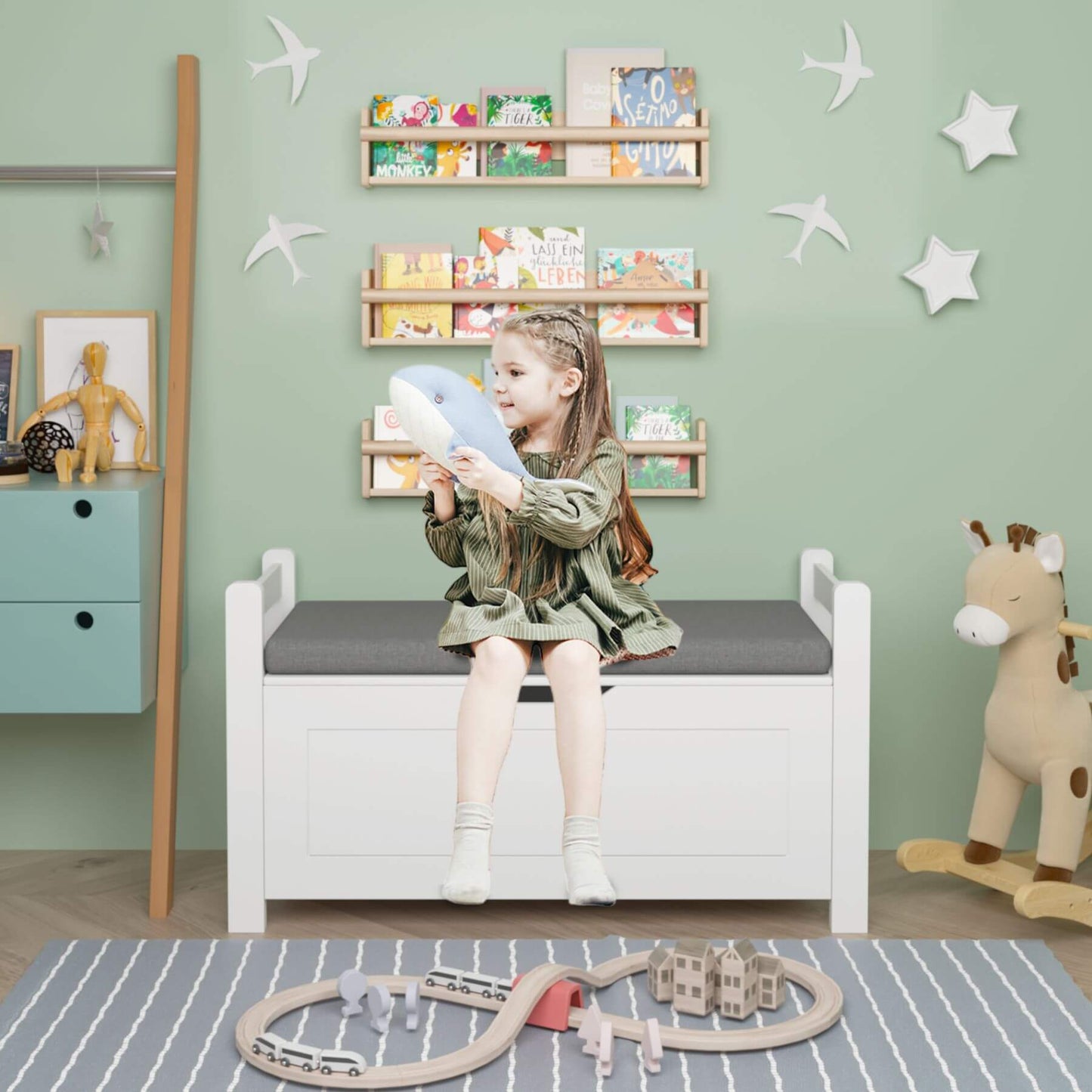 Child sitting on MOONRIVER Kids Toy Box Chest with a gray cushion, holding a blue toy, in a room with green walls, star decorations, and children's furniture.