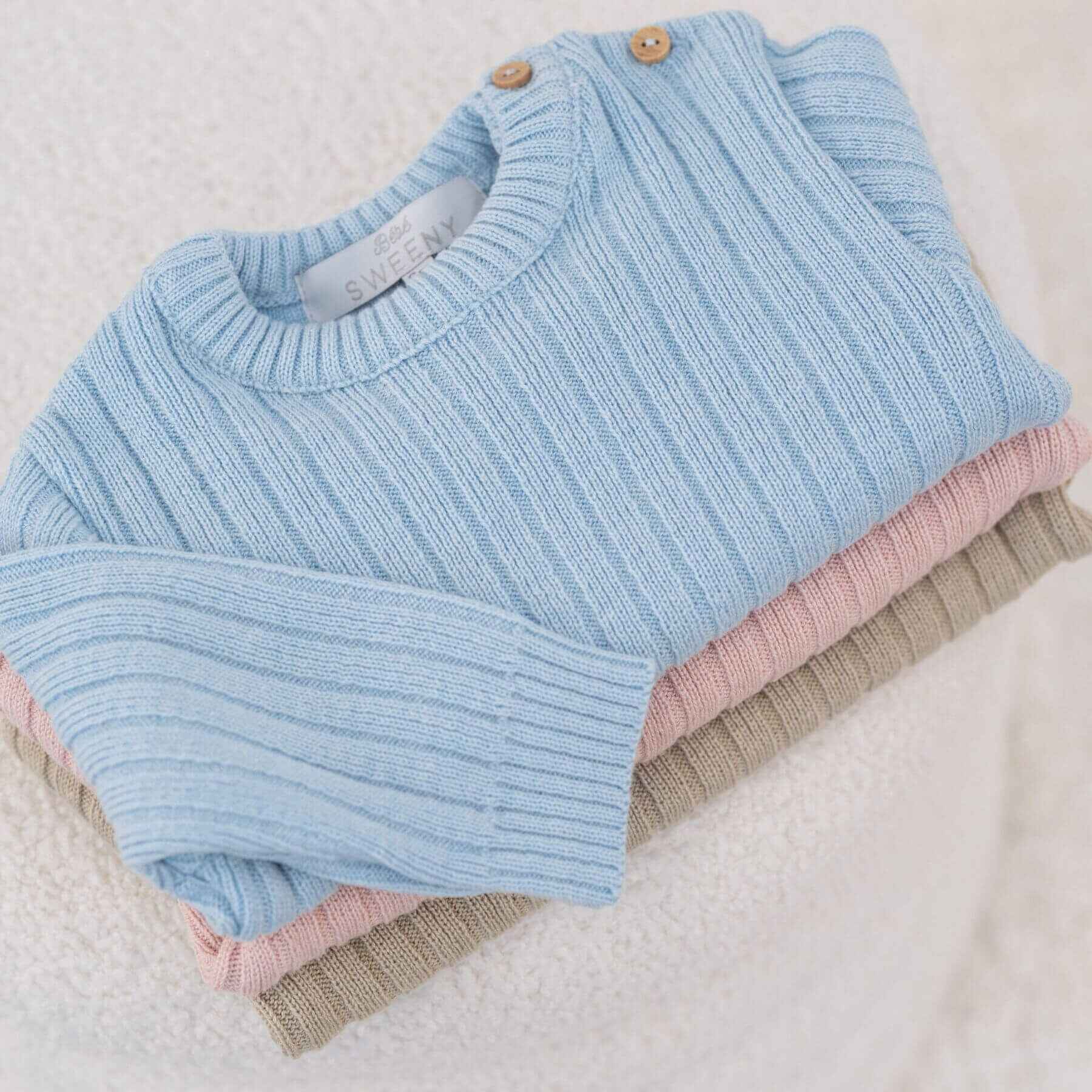 Stack of folded baby sweaters in blue, pink, and beige on a textured surface.