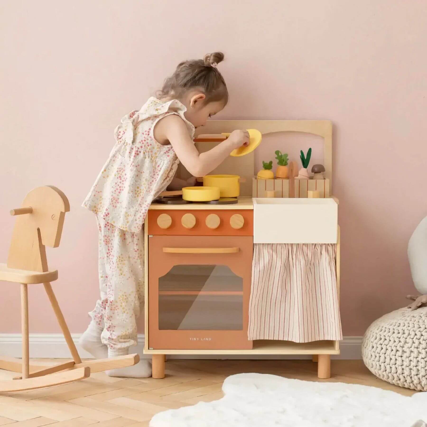 Child playing with Tiny Land® Cubby Kitchenette - Natural in a room with pink walls and wooden furniture.