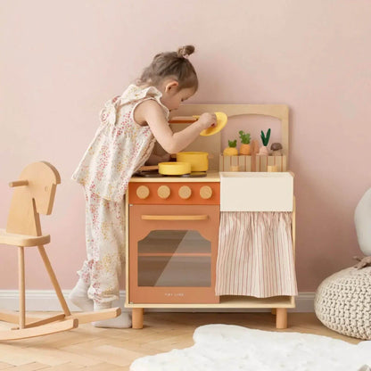 Child playing with Tiny Land® Cubby Kitchenette - Natural in a room with pink walls and wooden furniture.