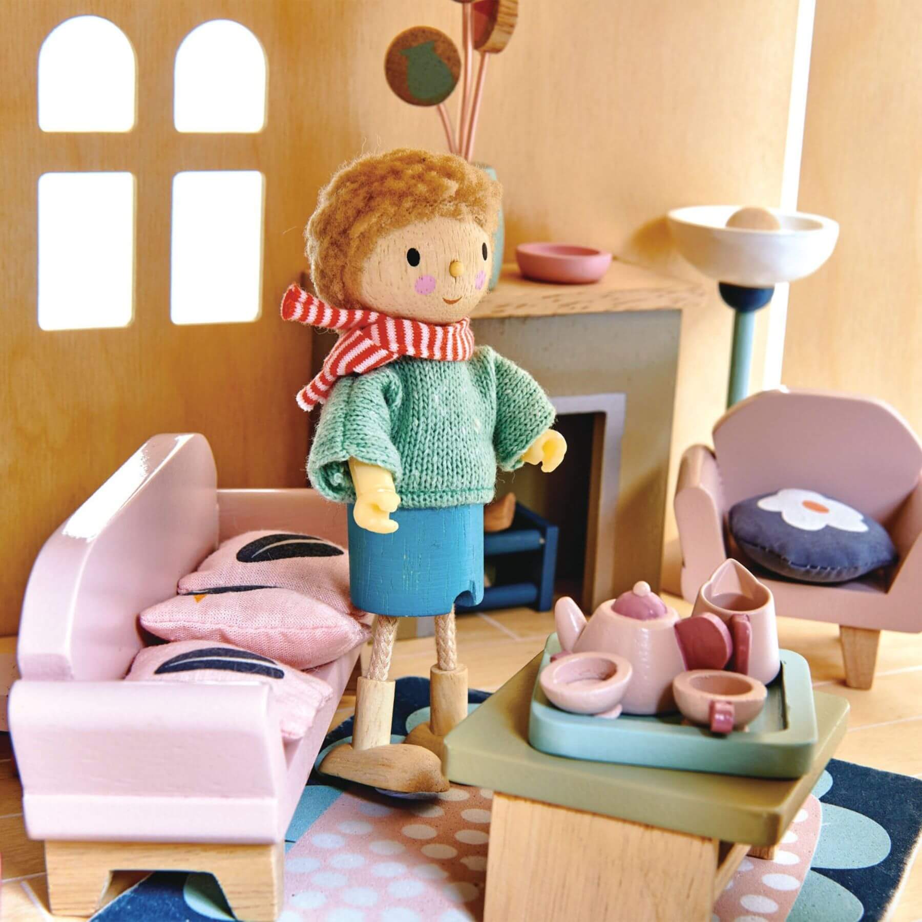 Doll in a green sweater standing in a wooden dollhouse living room with furniture and toys.