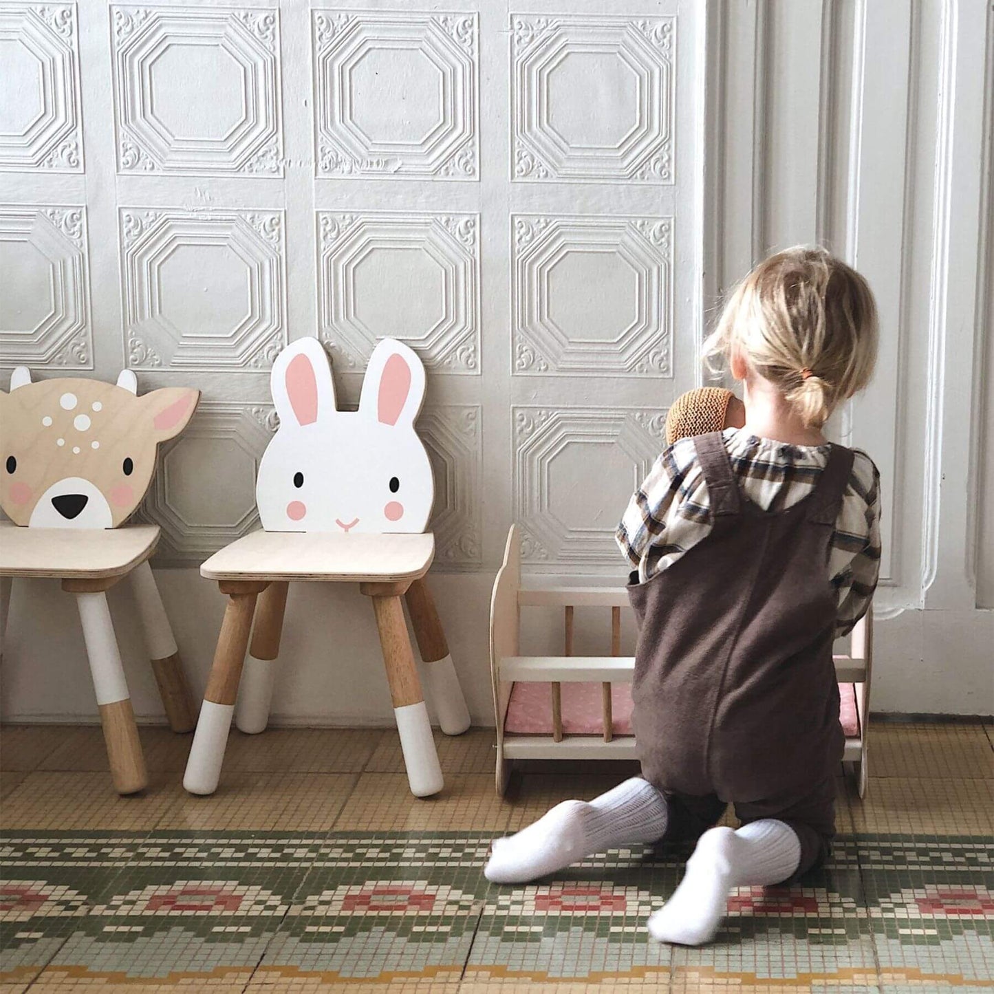 Child standing next to two wooden stools with animal faces in a room with decorative wallpaper.