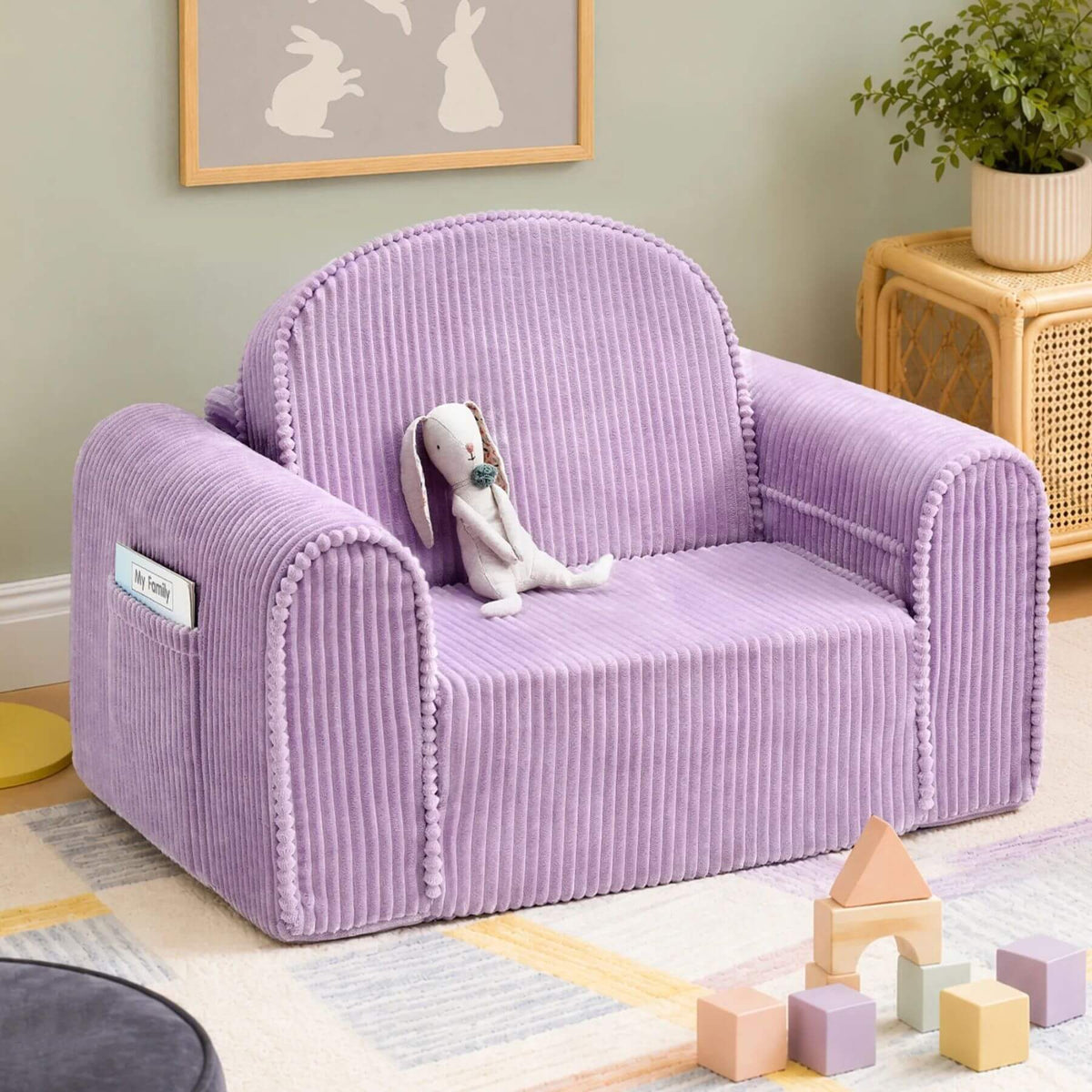 Tiny Land® GentleSnug Corduroy Kid Chair - Purple with a plush white rabbit toy resting on it, shown in a styled playroom setting with a rug, toy blocks, a side table, and framed wall art.