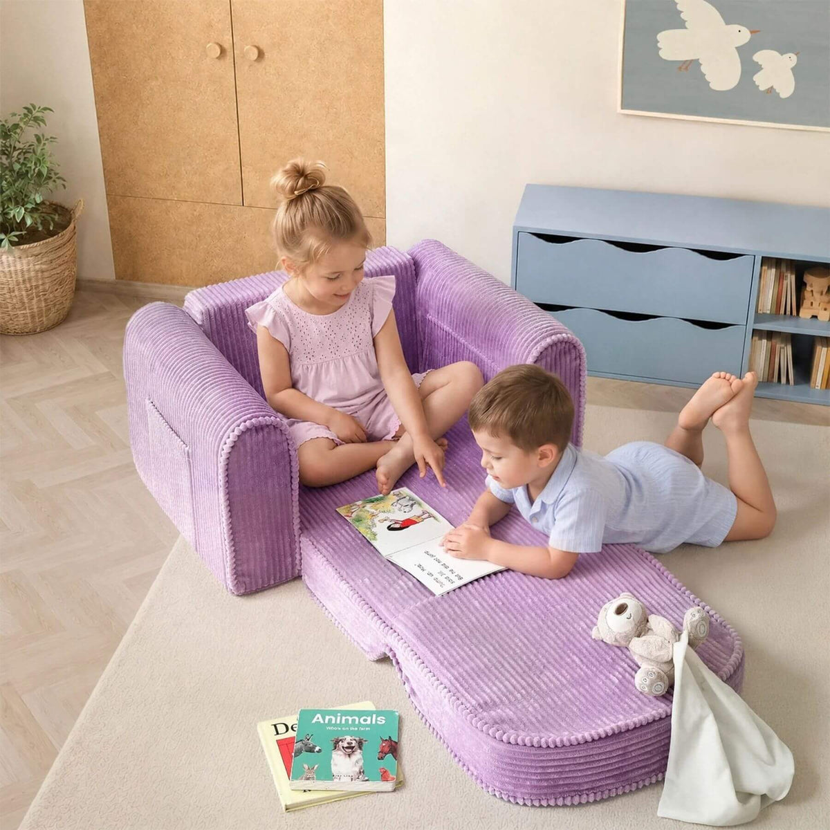 Tiny Land® GentleSnug Corduroy Kid Chair - Purple is set up on a light rug in a child's room. A girl sits on the armrests while a boy lies on the cushioned seat, reading a book.