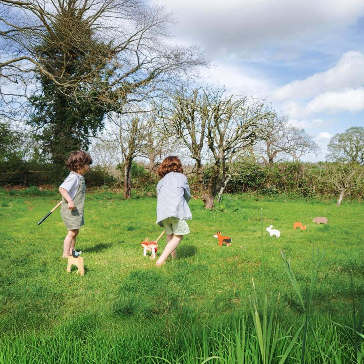 Two children playing with Tender Leaf Woodland Indoor Croquet Set in a grassy field on a sunny day.