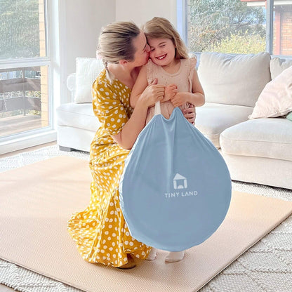 Woman in yellow polka dot dress holding a blue bag with 'Tiny Land' logo, child standing next to her in a living room.