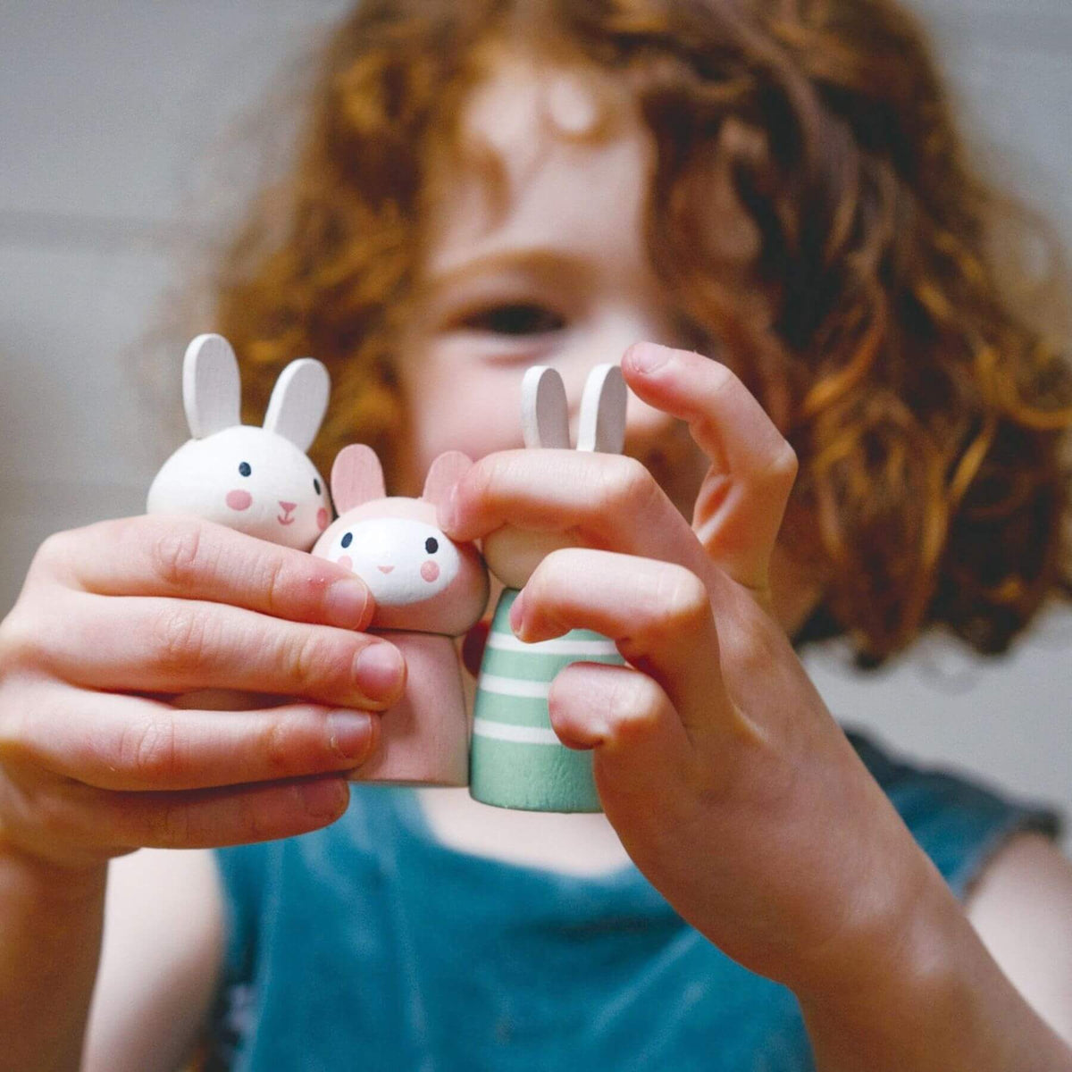 A close-up lifestyle shot of a young child with curly brown hair holding two small wooden bunny toys.