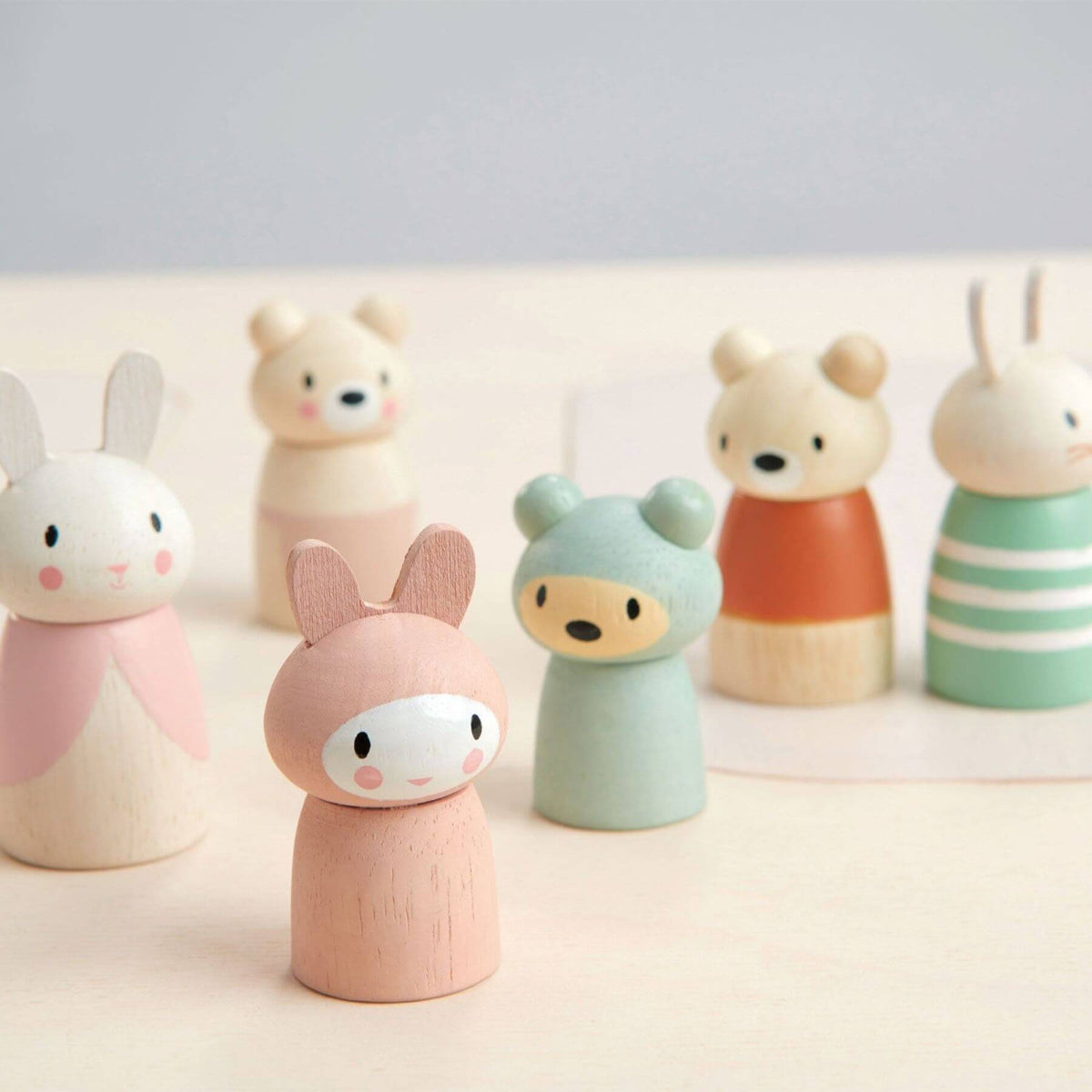 A set of small wooden animal figurines with pastel-colored painted bodies, including bunnies, bears, and a rabbit in a striped outfit, arranged on a light surface against a soft gray background.