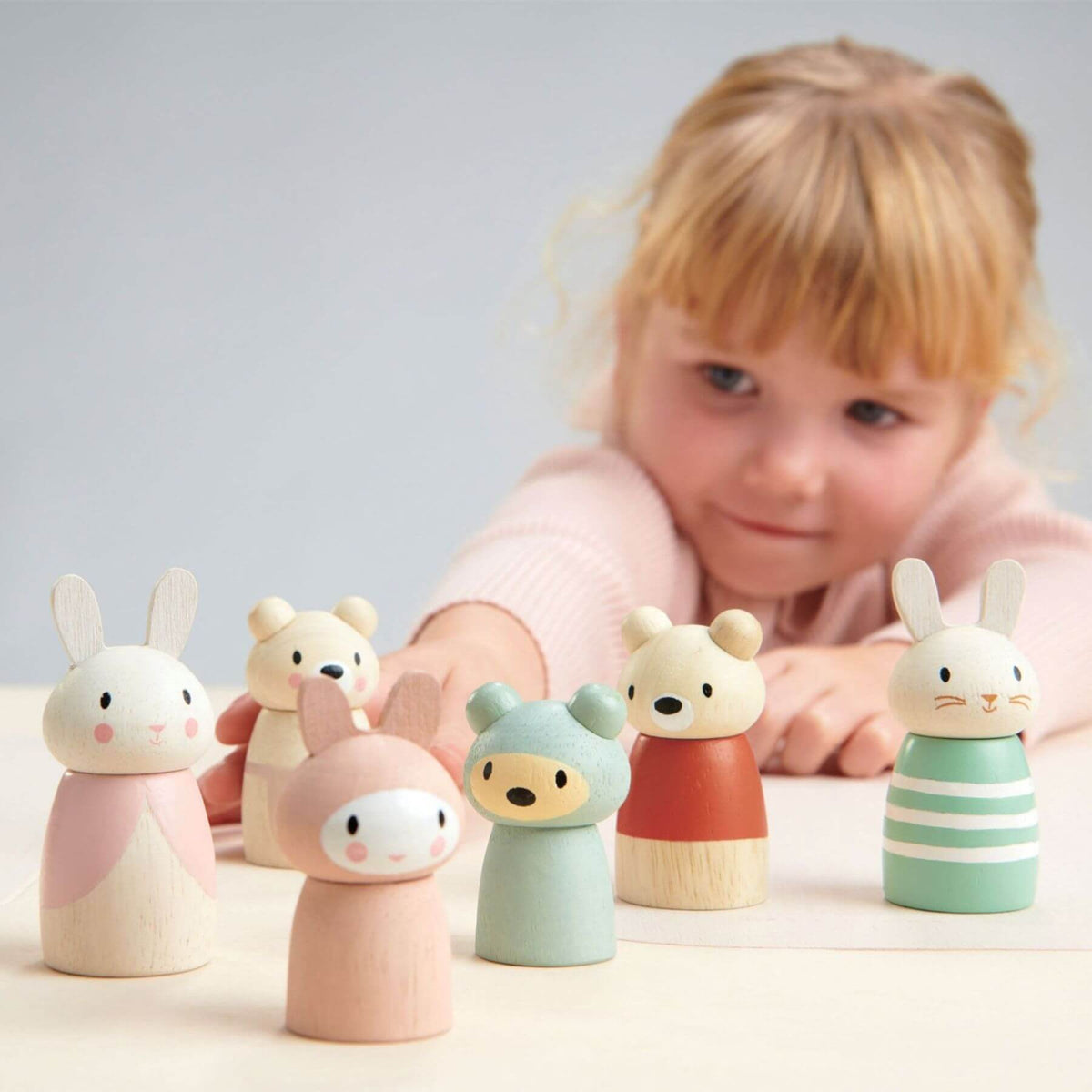 A young child with blonde hair leans over a table of pastel wooden animal toys, including bunnies, bears, and a bird figure, against a soft light gray background.