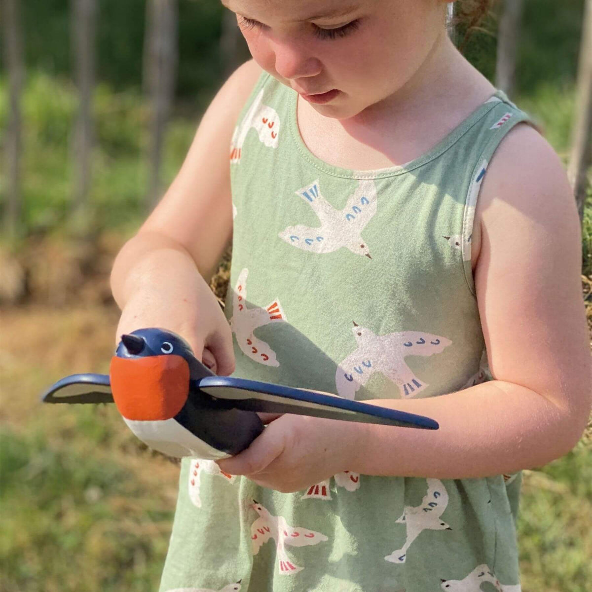 A young child outdoors holding a wooden toy bird. The child is wearing a sage green sleeveless dress with a white and blue bird pattern.