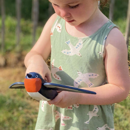 A young child outdoors holding a wooden toy bird. The child is wearing a sage green sleeveless dress with a white and blue bird pattern.