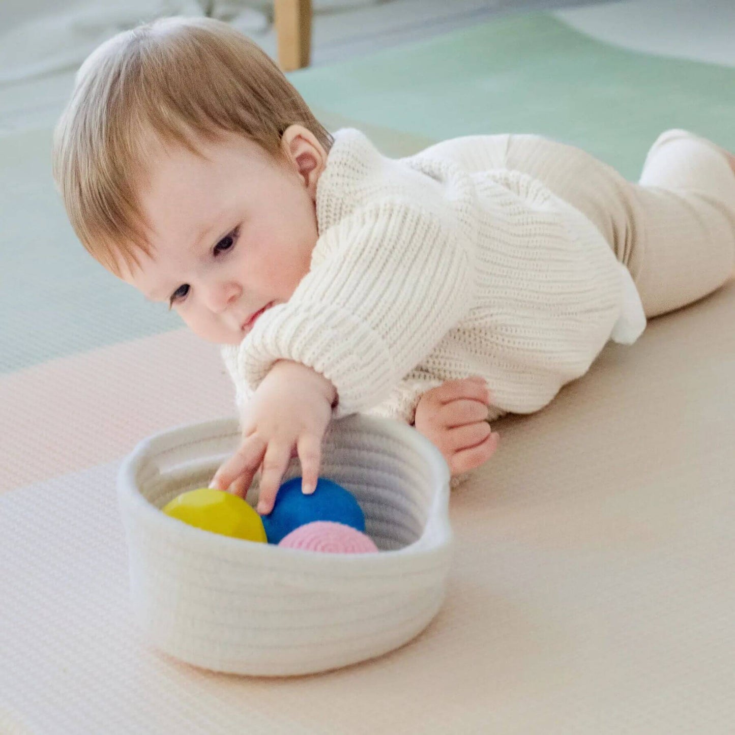 Baby playing with colorful balls in a basket on a light-colored floor.