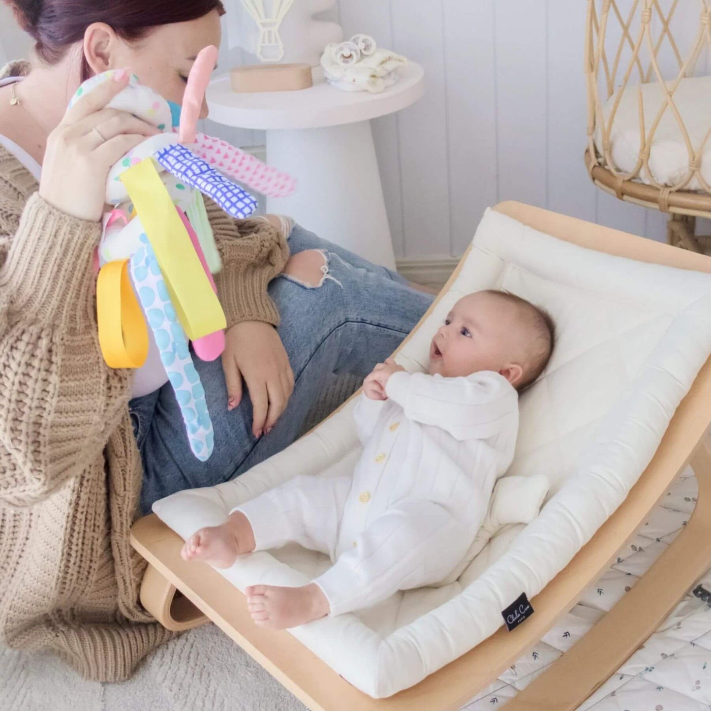 Baby in a rocker with a woman holding a colorful toy