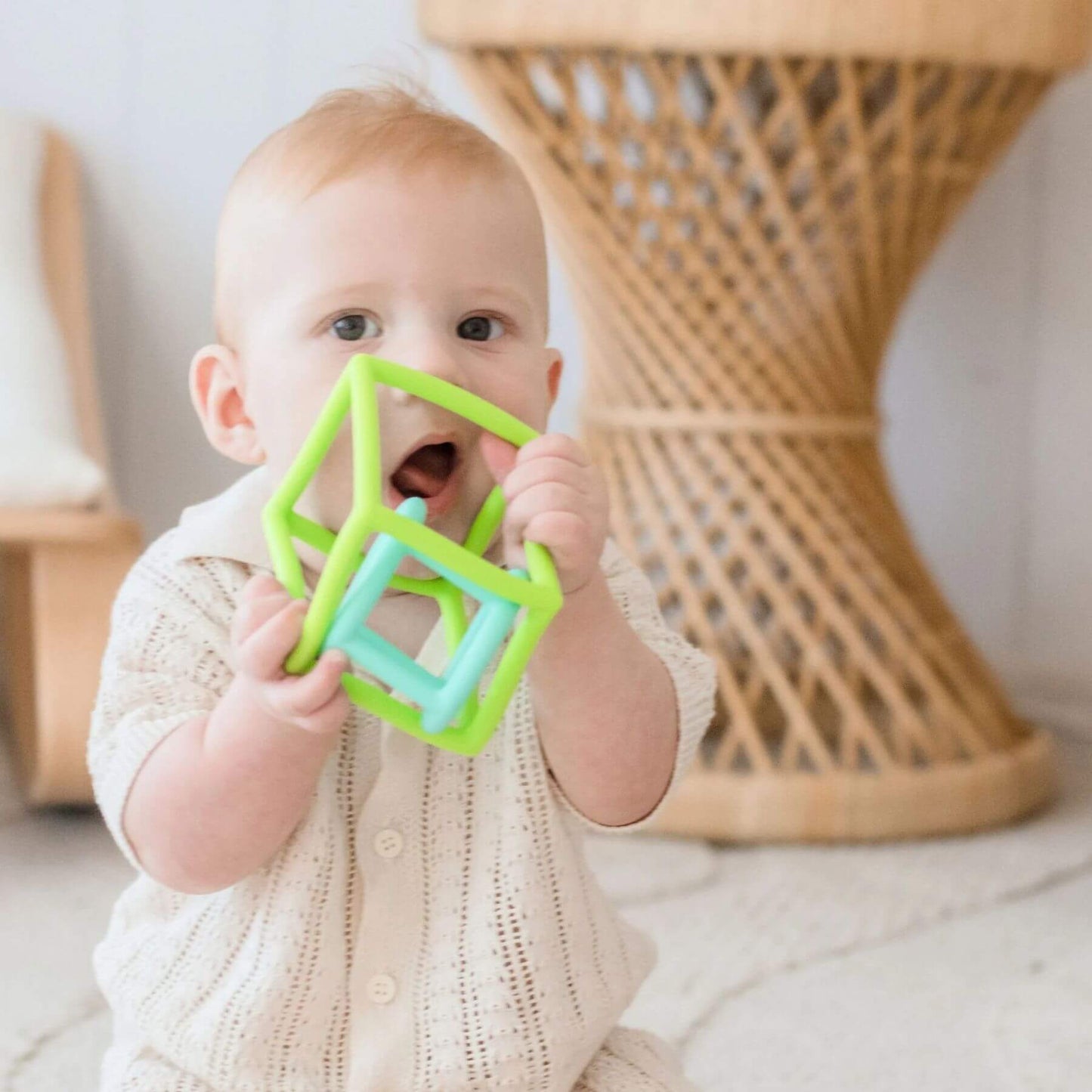 Baby holding a green and blue cube-shaped teething toy indoors.
