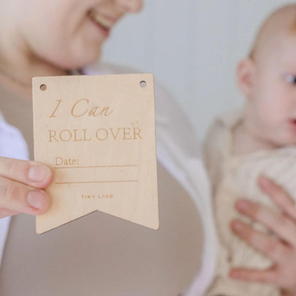 Person holding a wooden milestone tag with 'I Can Roll Over' text, blurred background