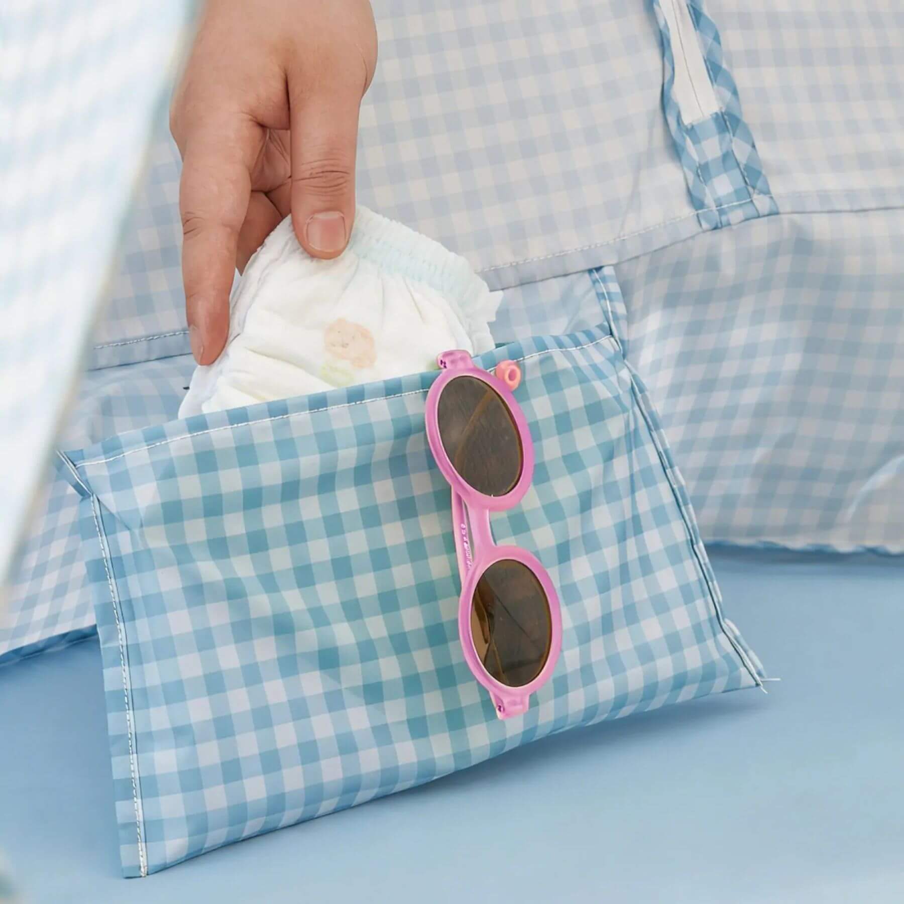 Hand reaching into a blue and white checkered bag with pink sunglasses and a diaper.