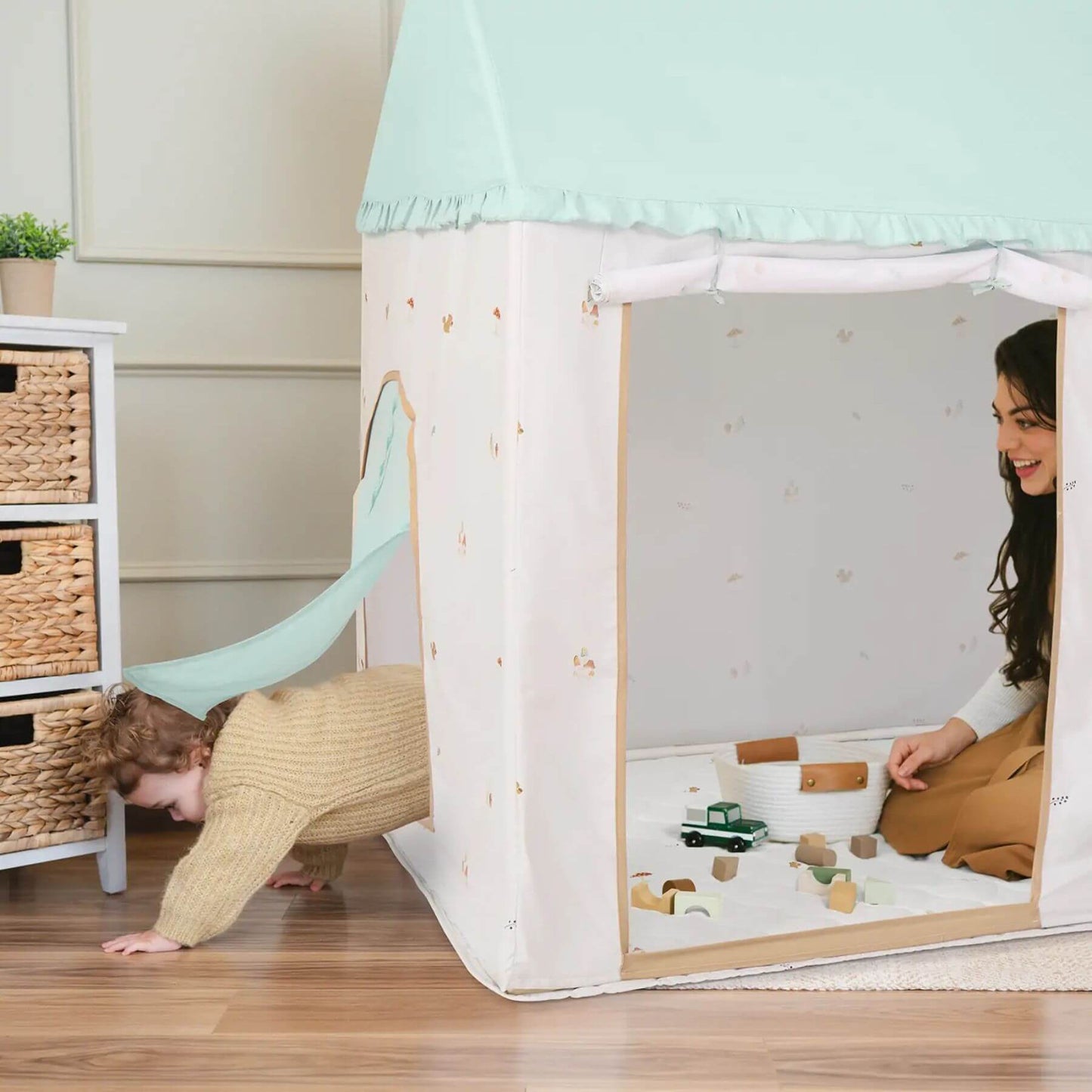 Child playing inside a play tent with a woman, in a room with a white dresser and wooden floor.