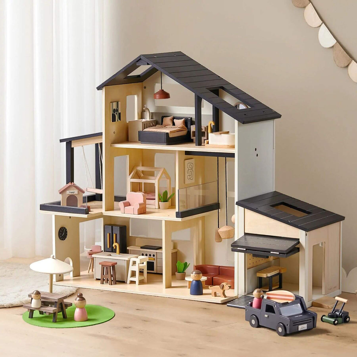 A large wooden dollhouse with an open roof and multiple interior rooms, shown on a light wood floor against a soft beige wall. The dollhouse includes a bedroom, kitchen, living area, garage, and small accessories like miniature furniture and toy vehicles.