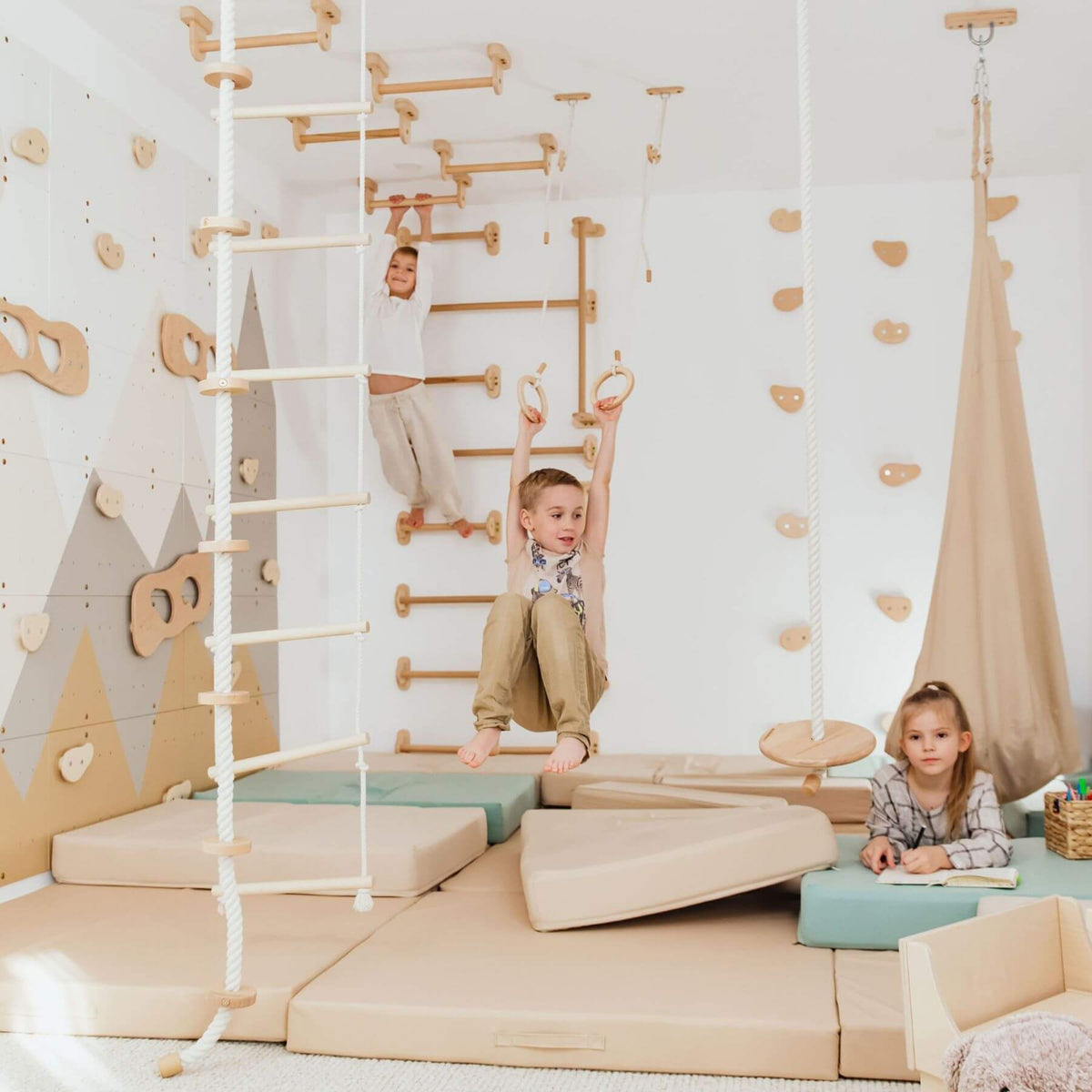 Wood and Hearts Climbing Wall Playground. Two children are actively playing in the space, with one boy hanging from wooden rings and another girl sitting at a table on the right.