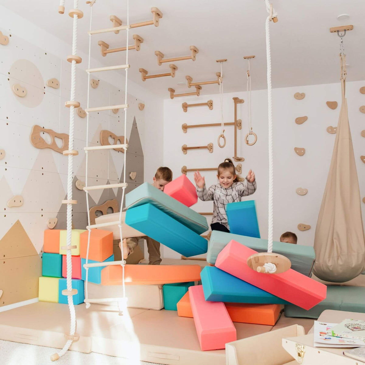 Children playing in a brightly lit indoor playroom with foam blocks, wooden climbing walls, rope ladders, and a hanging swing.