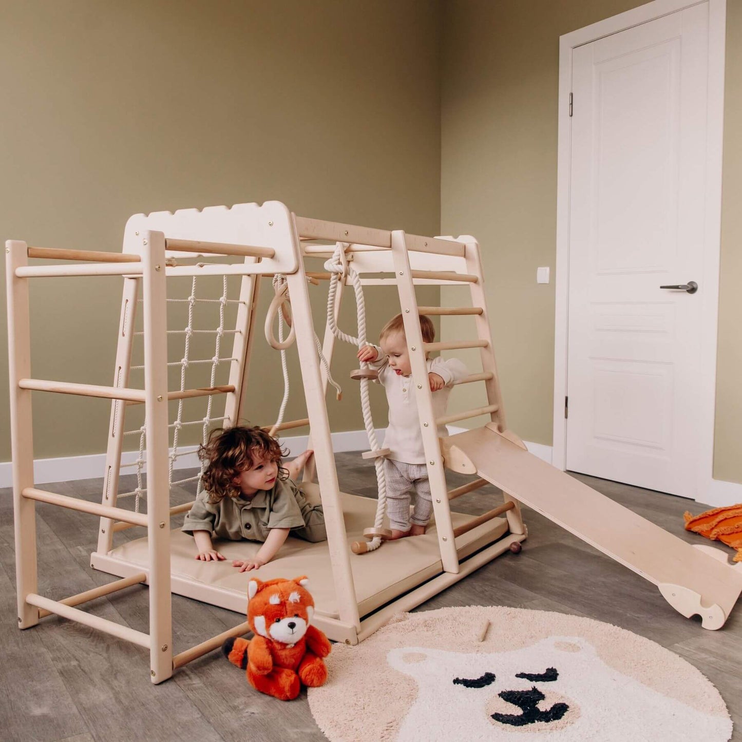 Children playing on Wood and Hearts Jungle Gym Playset with a teddy bear in a room.