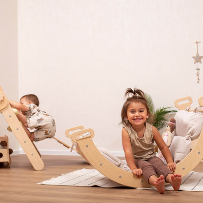 Two children playing on wooden toys in a room with a white wall and wooden floor.