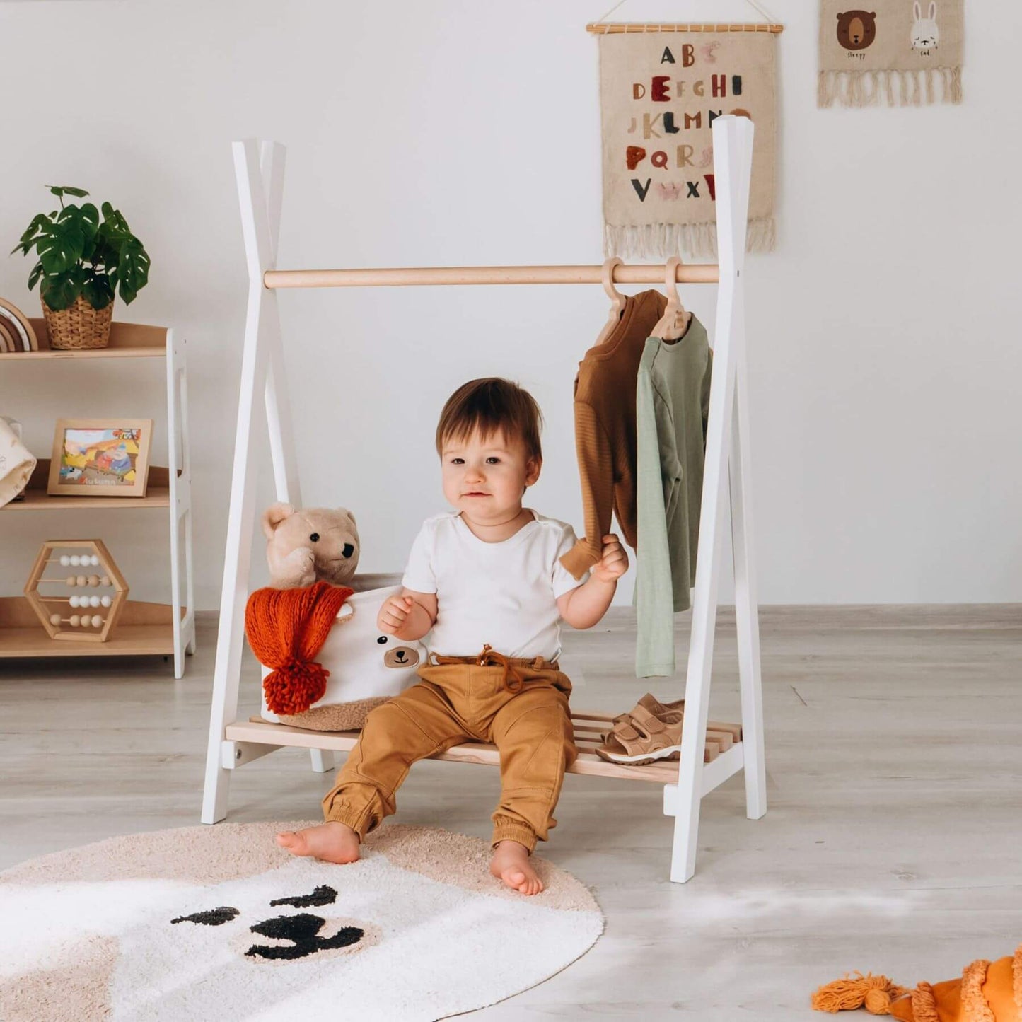 Child sitting on Wood and Hearts Toddler's Clothes Rack in a room with toys and a plant.