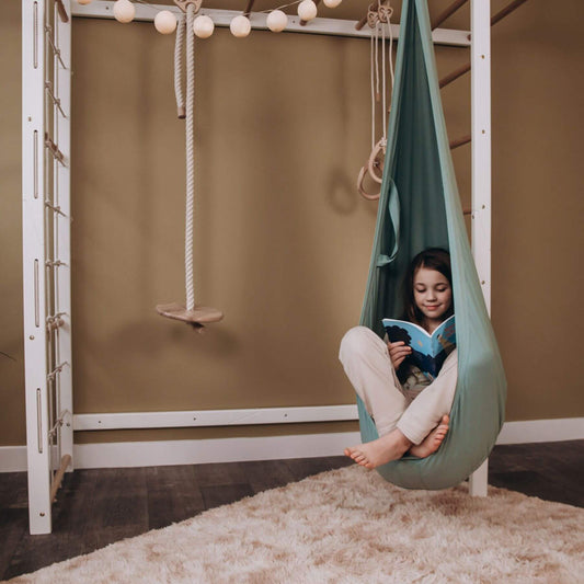 Child sitting in Wood and Hearts Pod Swing reading a book in a room with a ladder and swing.