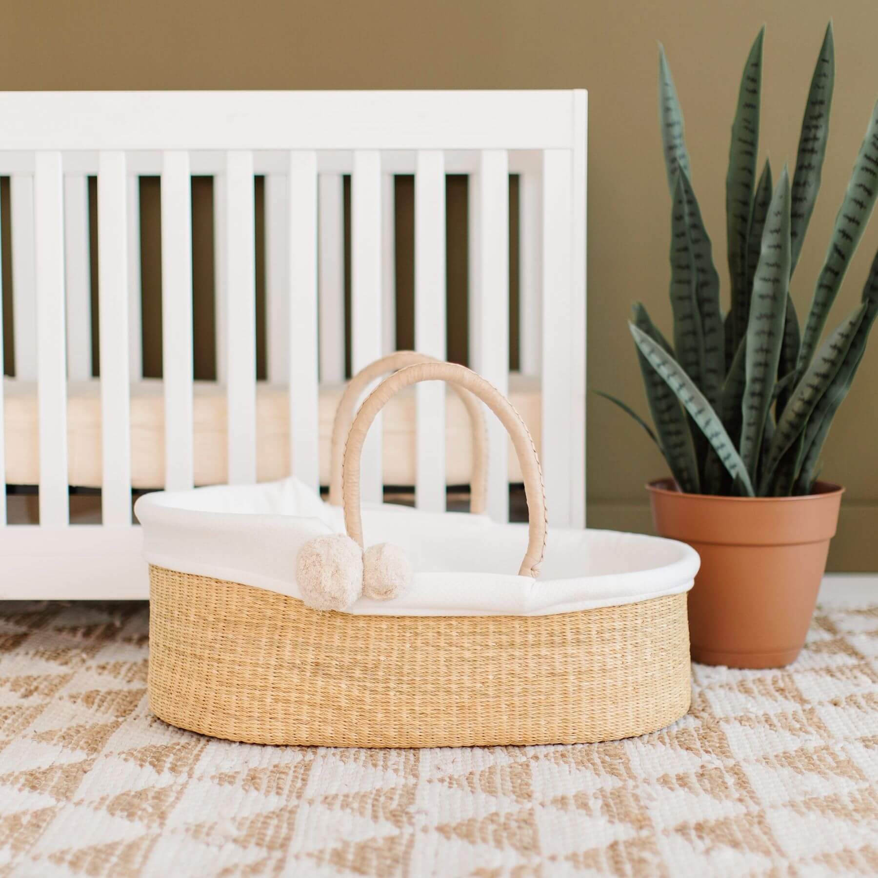 Plum+Sparrow African Moses Basket Cream Handle in front of a white crib with a plant in the background.