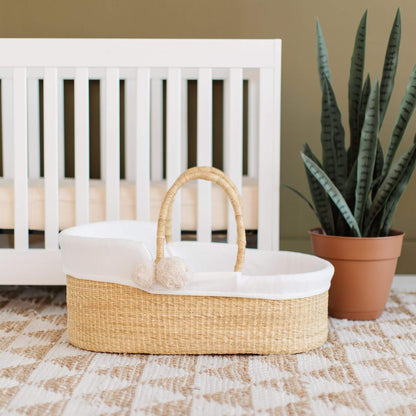 Plum+Sparrow Vegan Handle Moses Basket in front of a white crib with a potted plant on a patterned rug.