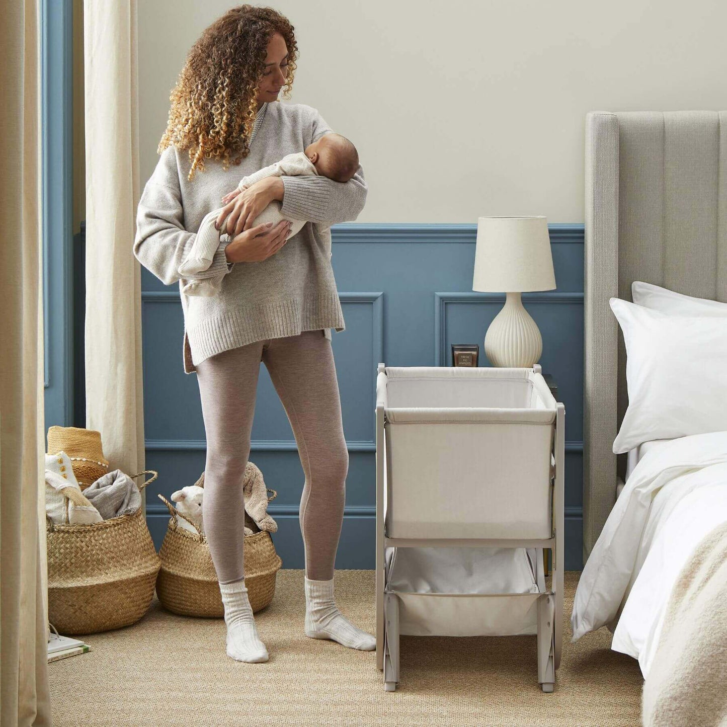 Woman holding a baby in a bedroom with Shnuggle Air Lite Bedside Crib.