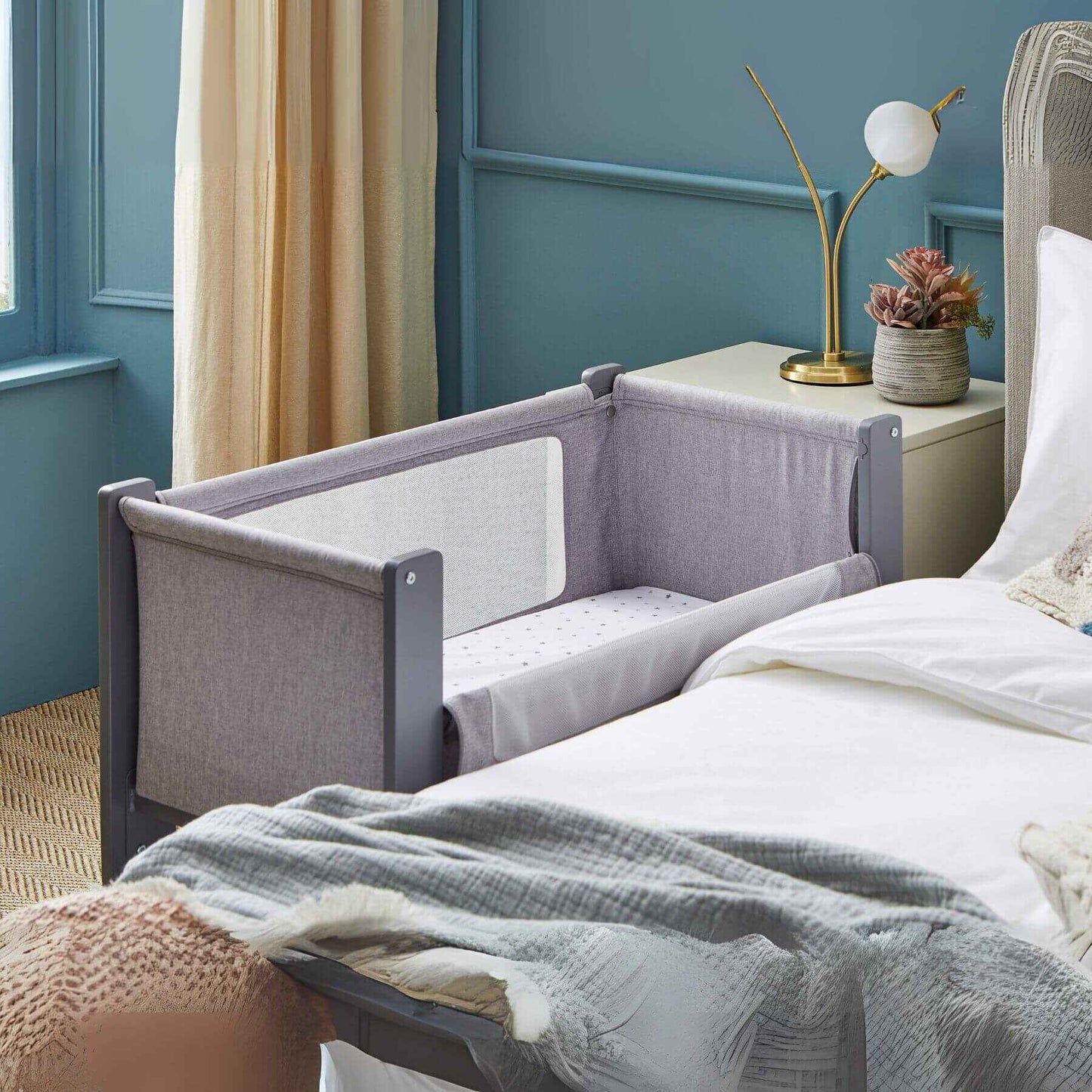 Shnuggle Air Lite Bedside Crib attached to a bed in a bedroom with blue walls and a lamp.