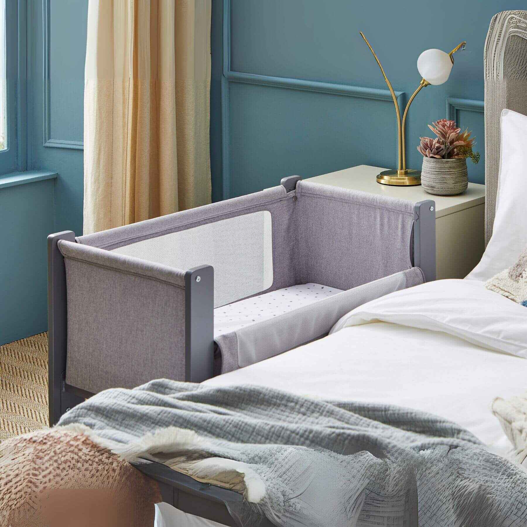 Shnuggle Air Lite Bedside Crib attached to a bed in a bedroom with blue walls and a lamp.