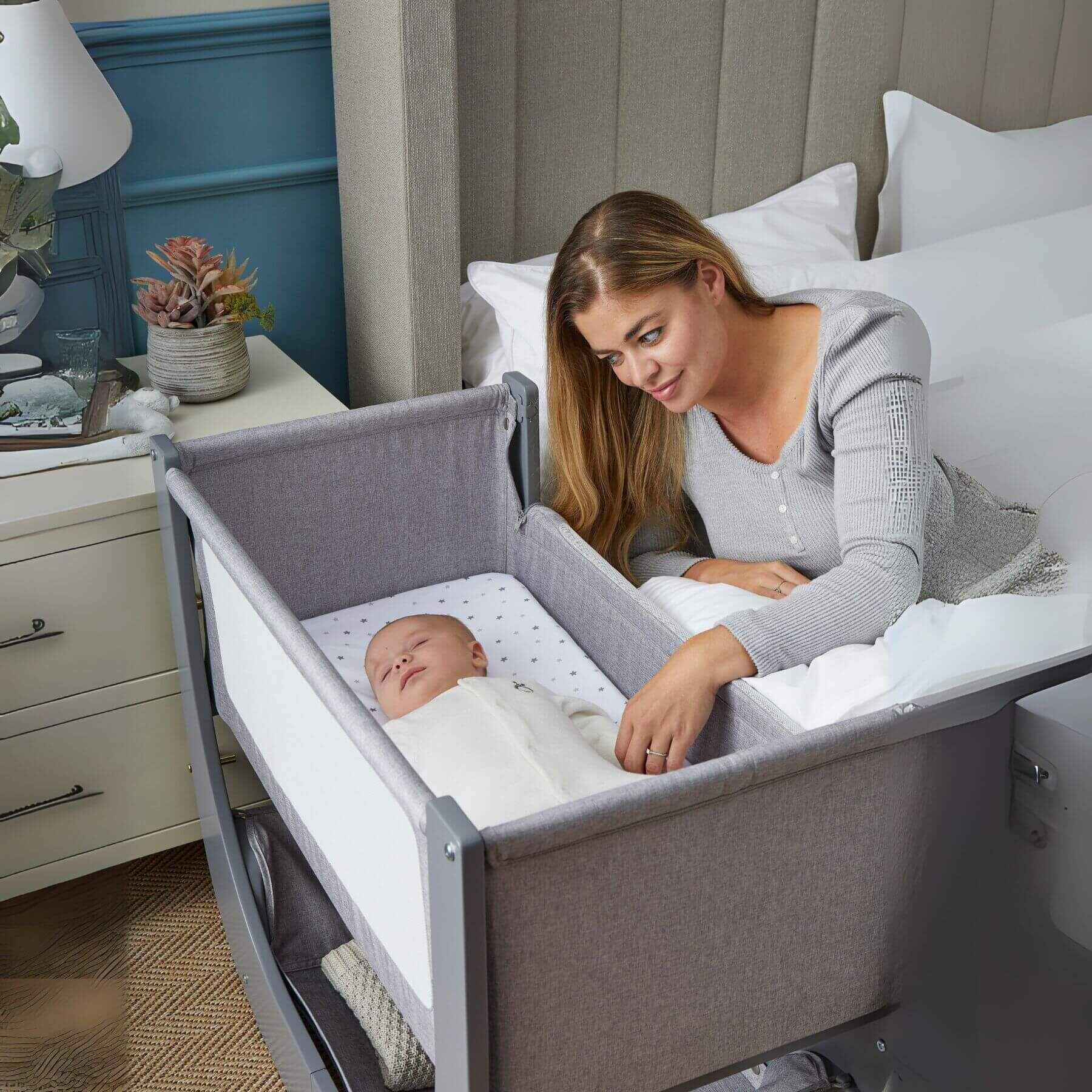 Woman lying on a bed next to a baby in Shnuggle Air Lite Bedside Crib in a bedroom setting.