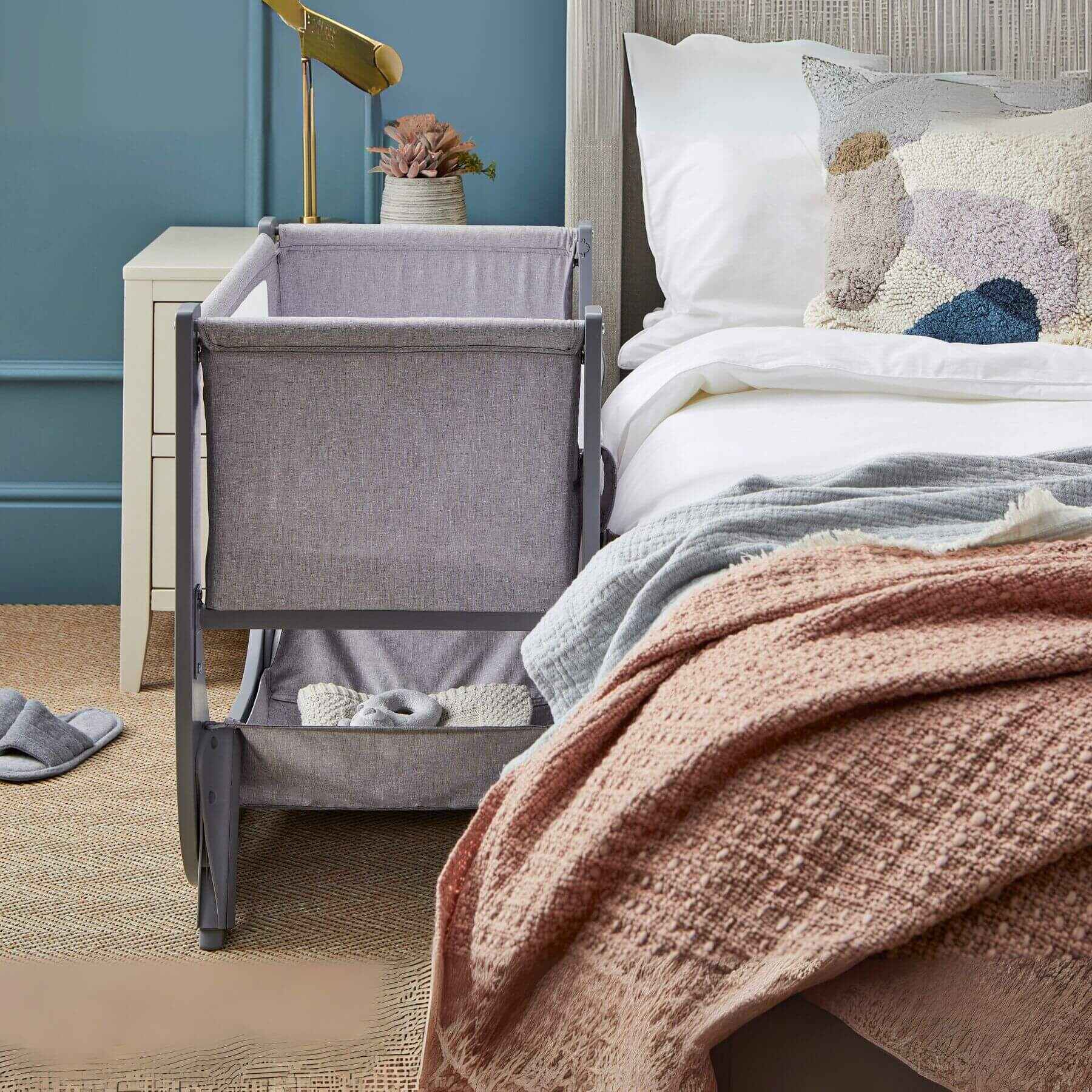 Shnuggle Air Lite Bedside Crib next to a bed with colorful pillows and a pink blanket in a bedroom setting.
