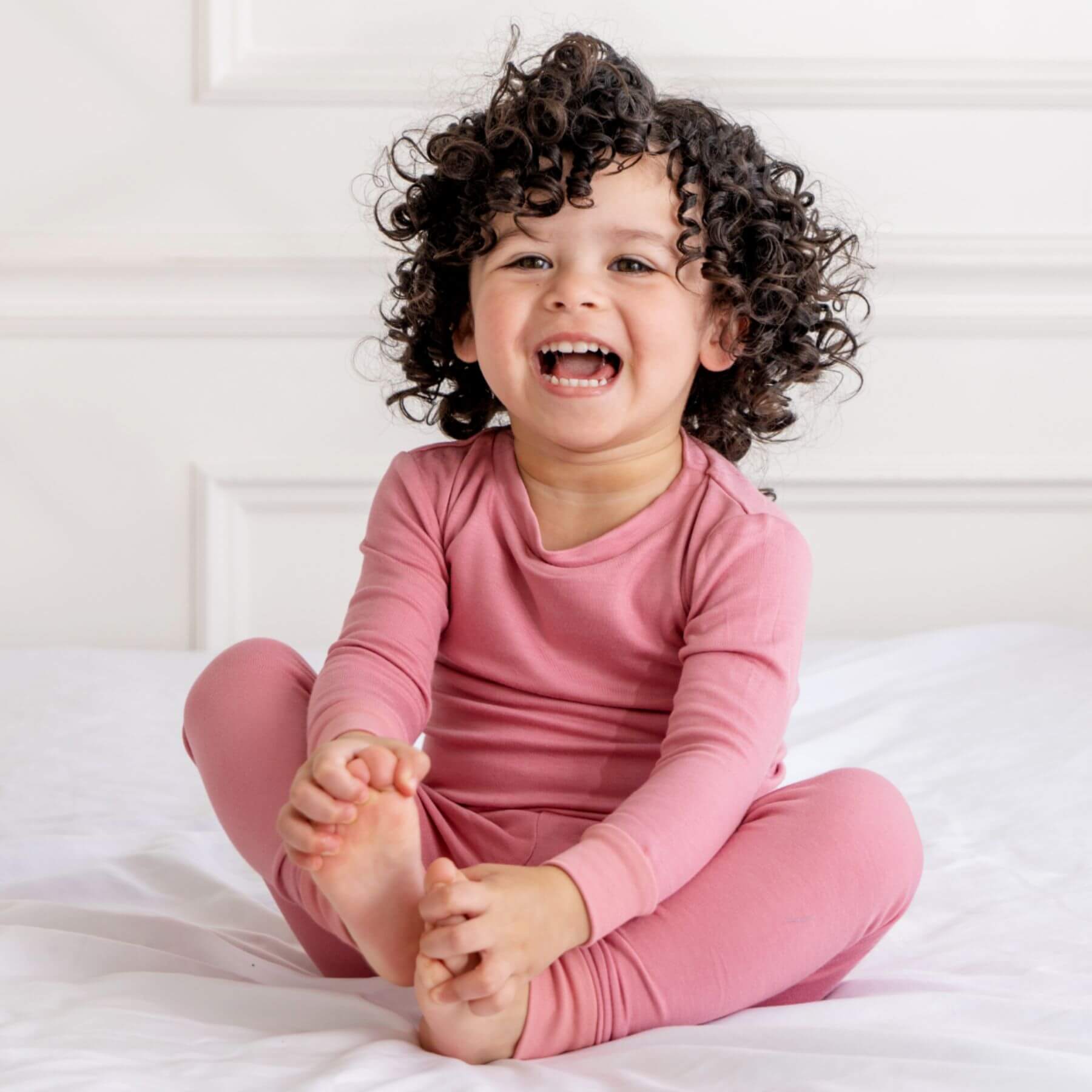 Child wearing Dreamland Baby Toddler/Kids Bamboo Pajamas - Dusty Rose sitting on a bed