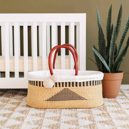 Plum+Sparrow Artisan Sky No Hood African Moses Basket on a checkered floor next to a white crib and potted plant.