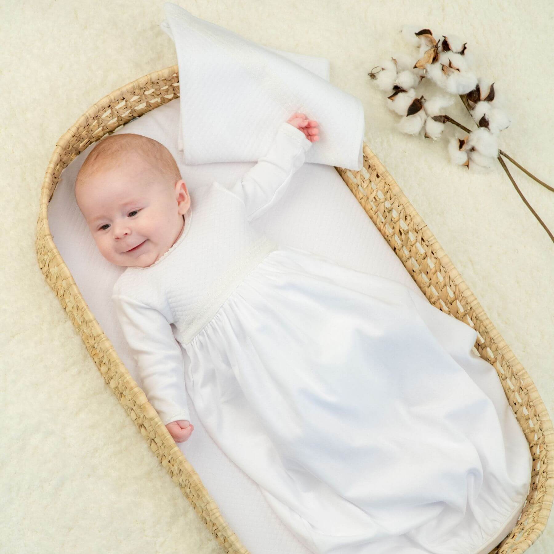Baby swaddled in Bebe Sweeny Baby White Quilted Cotton Gown lying in a woven basket on a beige surface with cotton flowers.