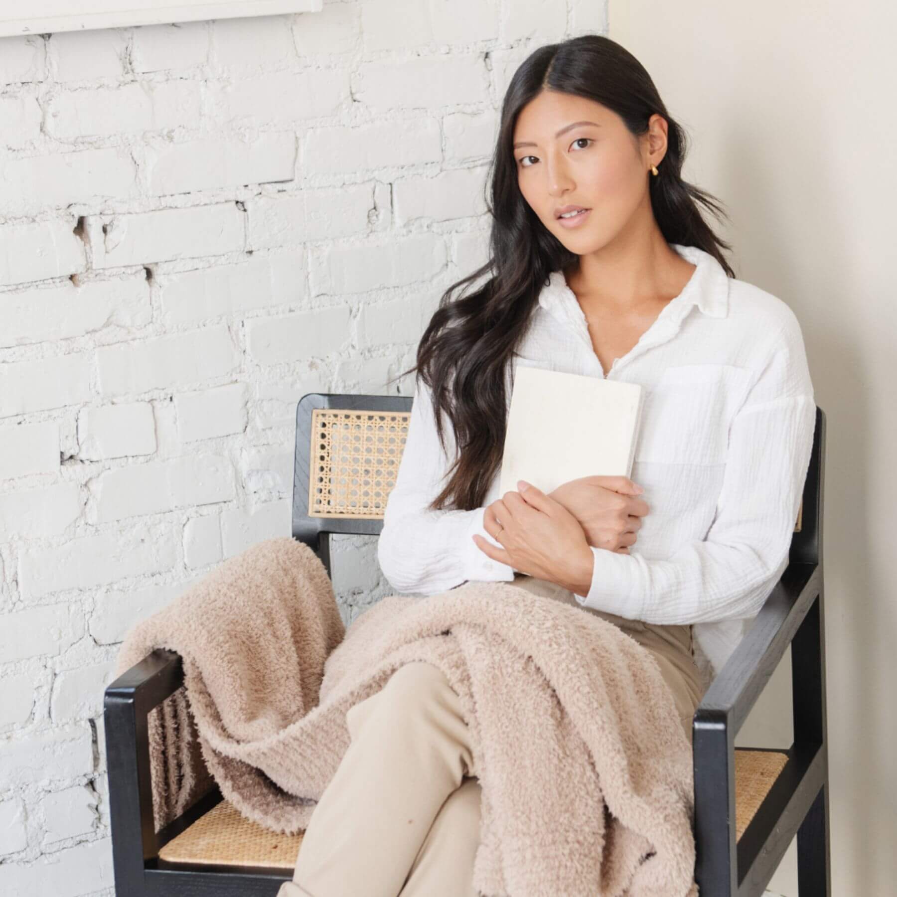Woman covered by Saranoni Bamboni® Throw Blanket, sitting on a chair holding a book against a white brick wall.