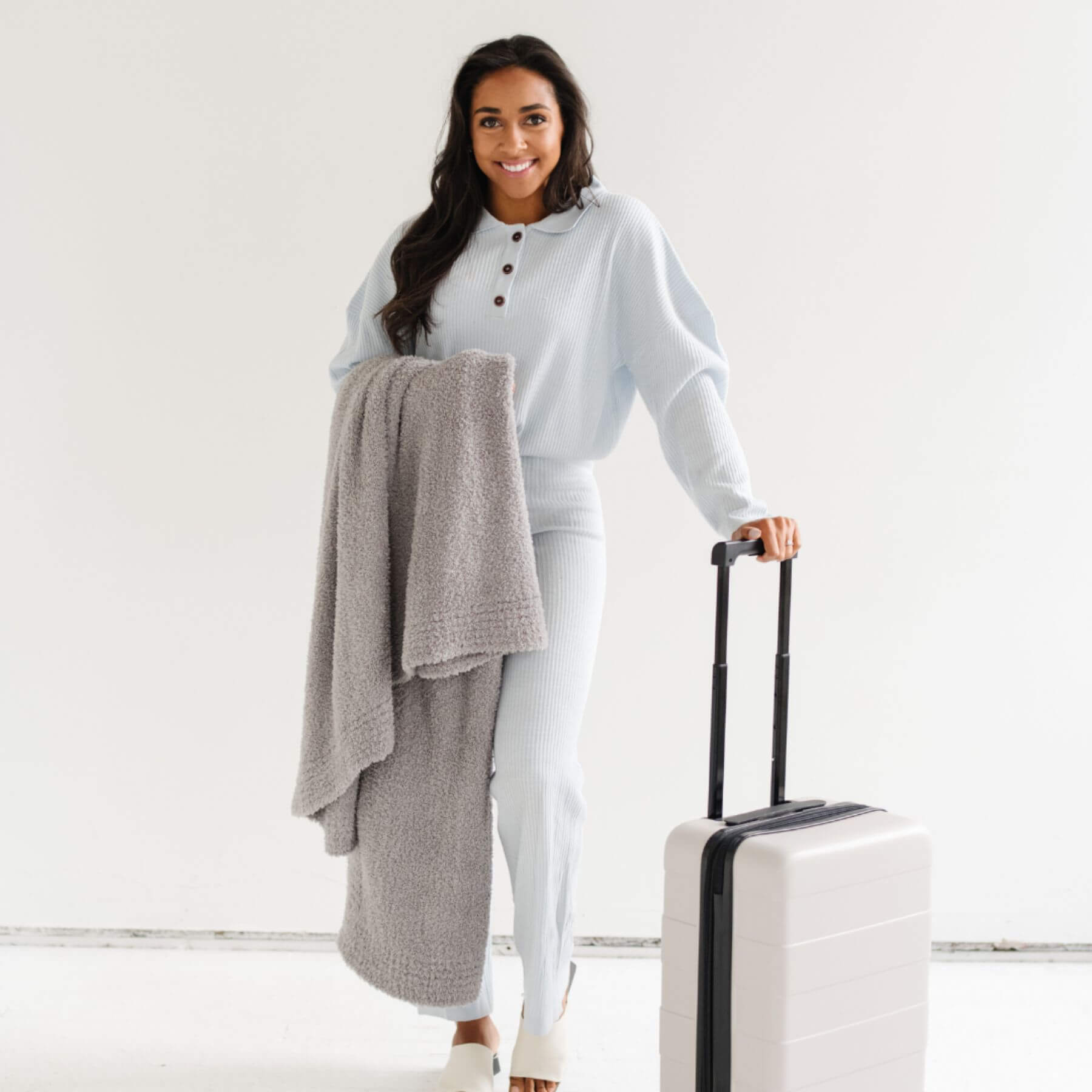 Woman holding Saranoni Bamboni® Throw Blanket, and a suitcase with a light gray outfit on a white background