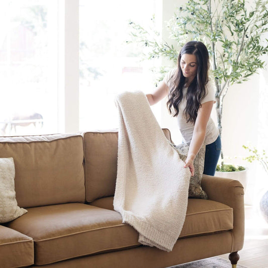 Woman arranging Saranoni Bamboni® Throw Blanket Taupe on a brown couch in a bright living room.